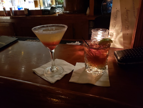 Two cocktails on a bar counter: a layered martini and a dark drink with a lime wedge, both on white napkins.