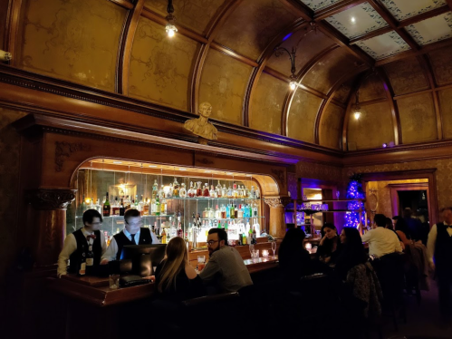 A stylish bar with a wooden interior, featuring bartenders serving drinks and patrons enjoying the ambiance.