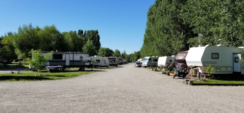 A gravel road lined with parked RVs and trailers, surrounded by green trees under a clear blue sky.