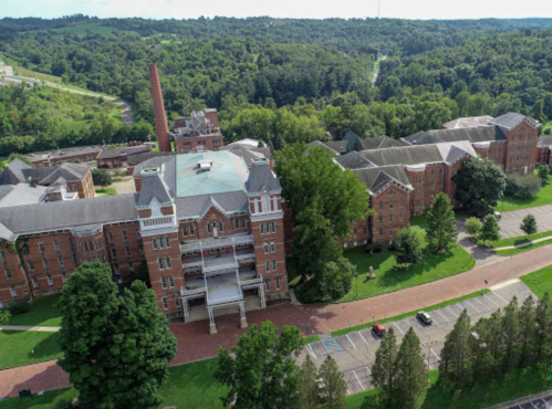 Aerial view of a large brick building surrounded by trees and a winding road in a green landscape.