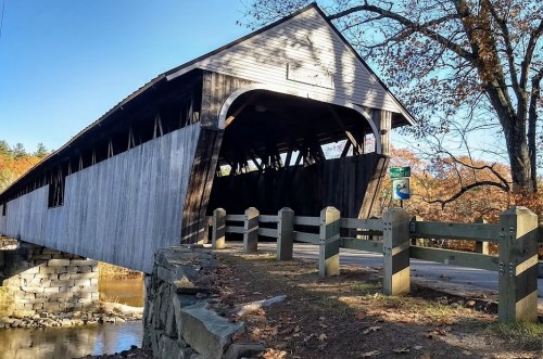 A wooden covered bridge spans a river, surrounded by autumn trees and a stone railing along the bank.