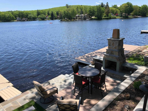 A serene lakeside view featuring a stone fireplace, outdoor seating, and lush green trees in the background.