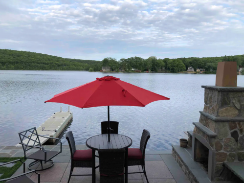 A red umbrella shades a table and chairs by a calm lake, surrounded by green trees and a cloudy sky.