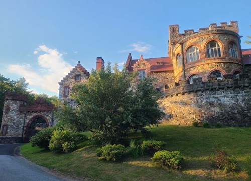 A historic stone castle with turrets, surrounded by greenery and a winding path under a clear blue sky.