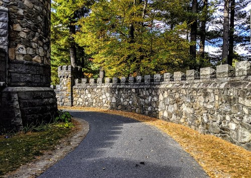 A winding path beside a stone wall, surrounded by trees with autumn foliage.