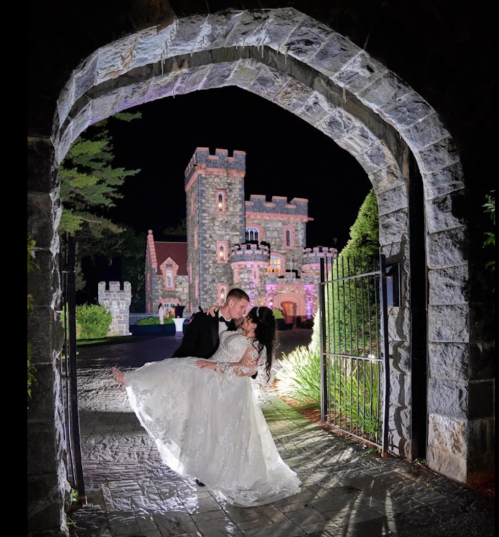 A couple in wedding attire poses romantically under a stone archway, with a castle illuminated in the background at night.