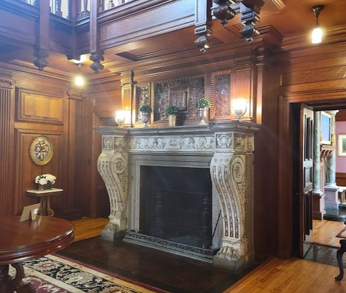 A richly decorated room featuring a large ornate fireplace and wooden paneling, with plants and soft lighting.