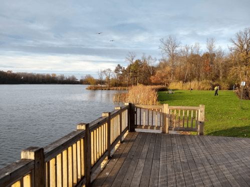 A serene lake scene with wooden decking, autumn foliage, and a person walking on the grassy shore.