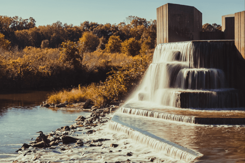 A serene waterfall cascades over a stone structure into a calm river, surrounded by autumn foliage.