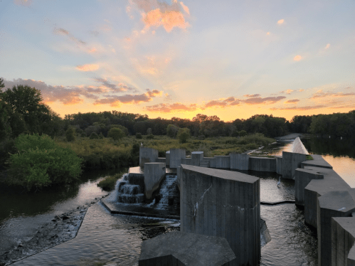 Sunset over a river with a modern concrete structure and small waterfalls, surrounded by lush greenery.