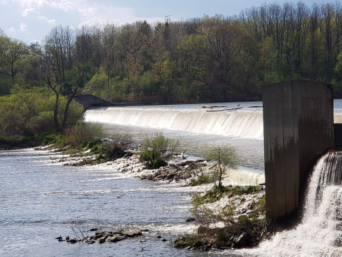 A serene river scene featuring a dam with cascading water, surrounded by lush greenery and trees in the background.