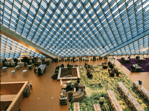 A spacious library interior with a unique glass ceiling, greenery, and people engaging in various activities.