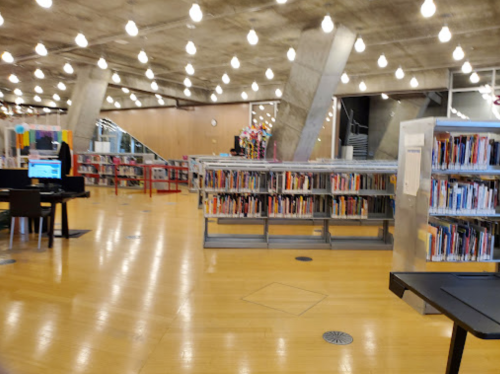 A modern library interior with wooden floors, bookshelves, and bright overhead lights.