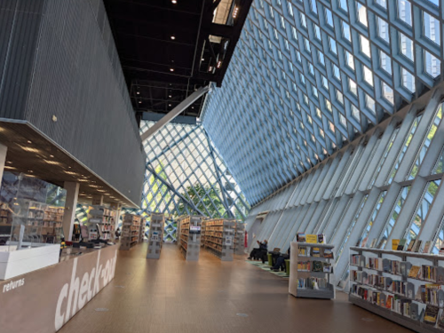 Modern library interior with large glass windows, wooden flooring, and shelves filled with books. Bright and spacious atmosphere.