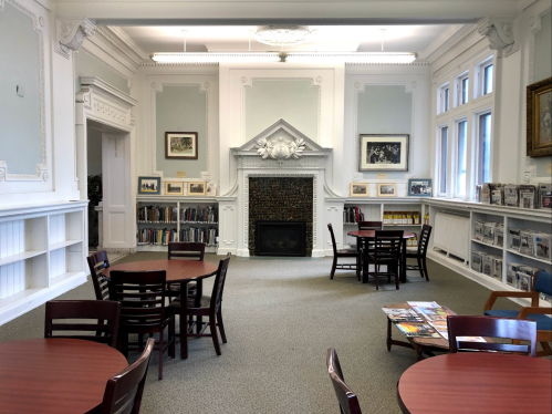 A spacious library room with tables, chairs, a fireplace, and bookshelves filled with books and photographs.
