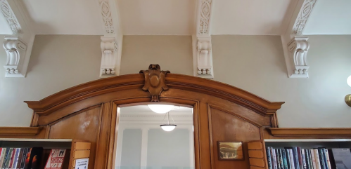 A wooden archway with ornate detailing above, leading to a room lined with bookshelves. Elegant ceiling features visible.