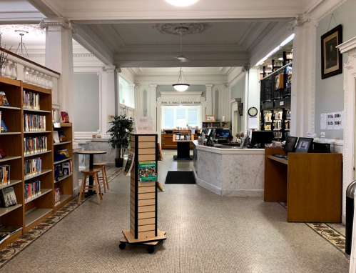 Interior of a library featuring bookshelves, a circulation desk, and a display stand on a tiled floor.
