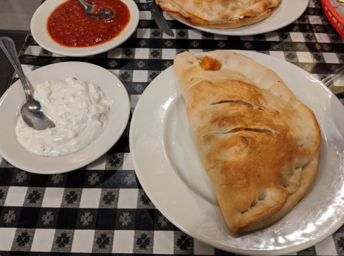 A calzone on a plate next to a bowl of white sauce and a small dish of red sauce, all on a checkered tablecloth.