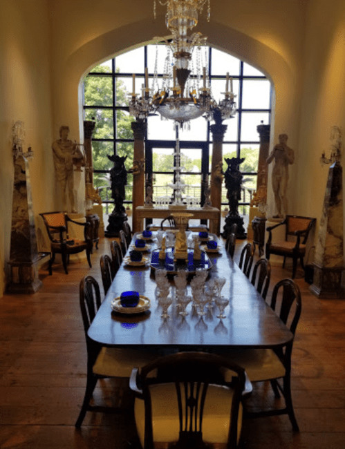 Elegant dining room featuring a long wooden table, ornate chandelier, and classical sculptures by large windows.