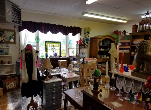 A cozy antique shop filled with vintage furniture, glassware, and decorative items, illuminated by natural light.