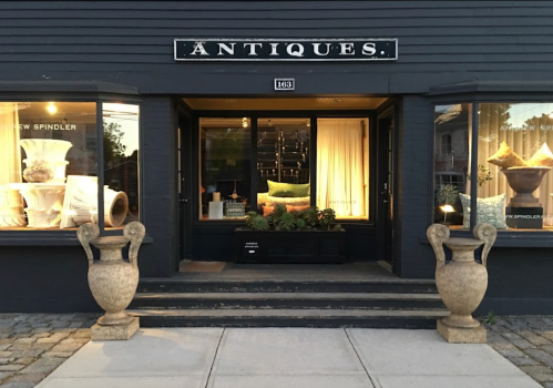 A storefront labeled "Antiques" with large windows displaying decorative items and two stone urns at the entrance.