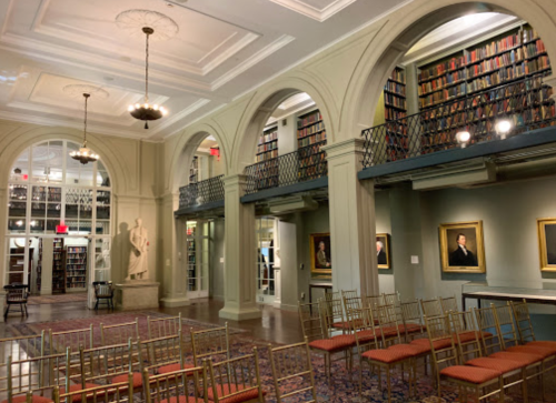 A spacious library with tall bookshelves, elegant arches, and rows of chairs set up for an event.