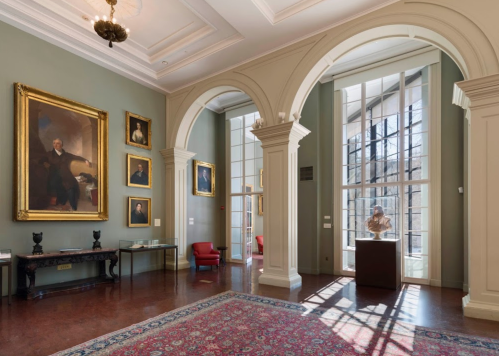 A spacious gallery with arched doorways, large windows, paintings on the walls, and a sculpture on a pedestal.