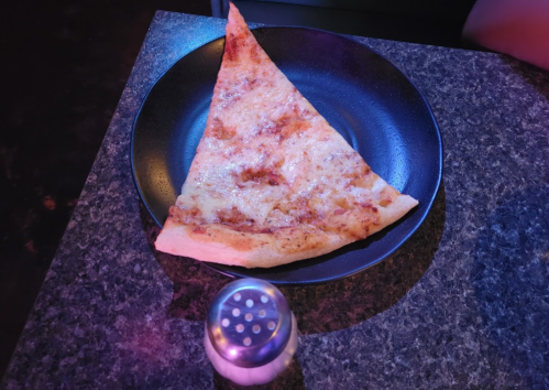A large slice of cheese pizza on a black plate, next to a salt shaker, set against a dark background.