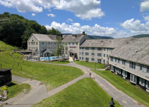 A resort building with a pool, surrounded by green grass and trees under a partly cloudy sky.