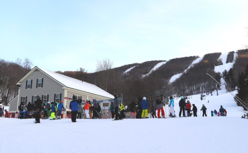 A busy ski resort with people in colorful gear waiting in line near a lodge, surrounded by snowy slopes and trees.