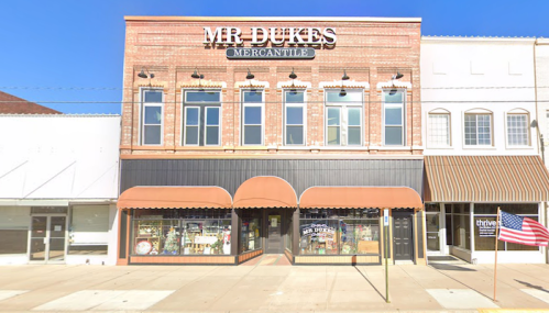 A brick building with "Mr. Dukes Mercantile" sign, featuring large windows and awnings, on a sunny street.