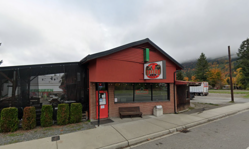 A red building with a sign, featuring a bench outside, surrounded by trees and a cloudy sky.