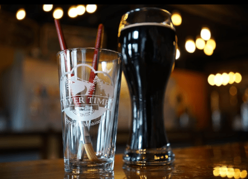 A clear glass with straws and a dark beer in a tall glass, set on a wooden bar with warm lighting in the background.