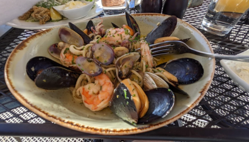 A bowl of seafood pasta featuring shrimp, mussels, clams, and squid, garnished with herbs on a textured plate.