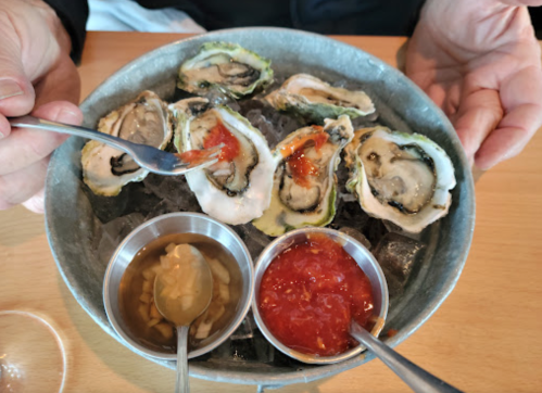 A person holds a metal platter of oysters on ice, accompanied by two sauces in small bowls.