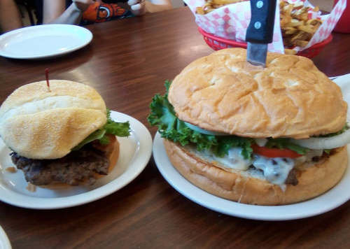A small burger with lettuce and a toothpick next to a large burger topped with lettuce, tomato, and a knife.