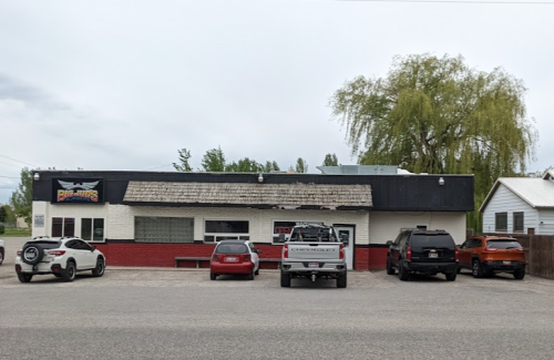 A small, one-story building with a black roof and red accents, featuring parked cars in front.