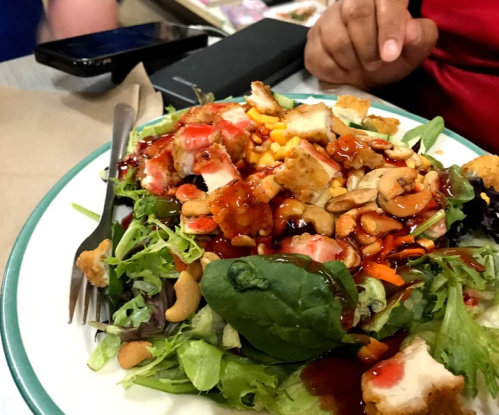A colorful salad topped with grilled chicken, nuts, and a sweet sauce, served on a white plate with a fork.