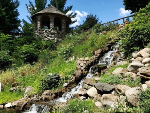 A stone gazebo overlooks a lush hillside with a cascading waterfall and vibrant greenery.