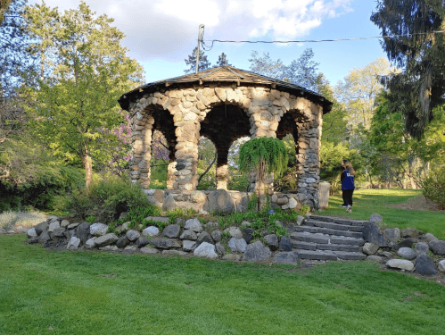 A stone gazebo surrounded by greenery, with a person walking nearby on a grassy path.