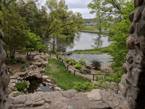 View of a serene pond surrounded by greenery, framed by stone walls and trees in a peaceful outdoor setting.