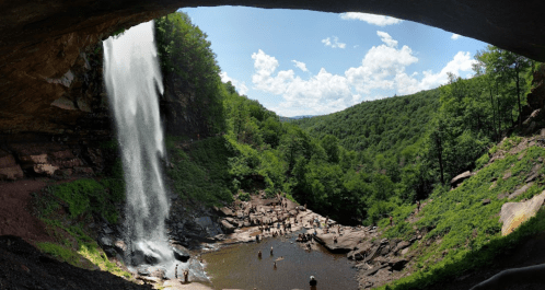 A panoramic view of a waterfall cascading into a rocky area, surrounded by lush green hills and a blue sky.