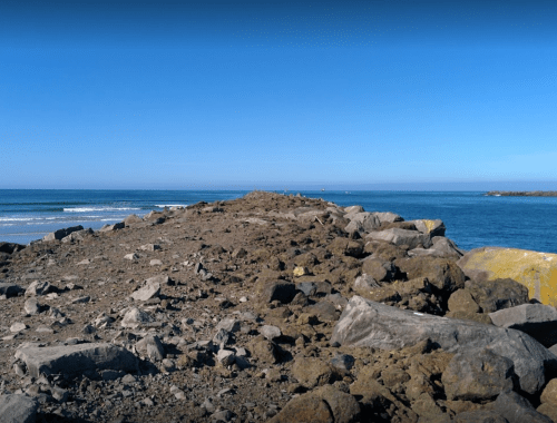 A rocky jetty extends into a calm blue sea under a clear sky, with gentle waves lapping at the shore.