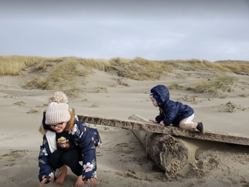 Two children play on a sandy beach; one crouches down while the other climbs on a log in the background.