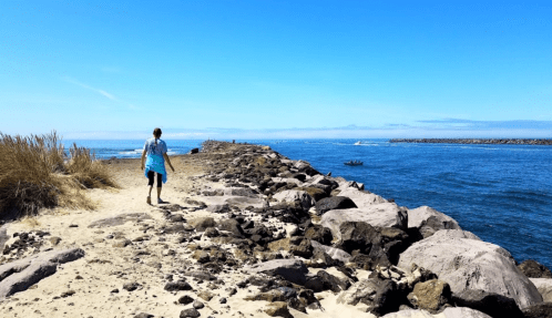 A person walks along a rocky path by the water, with blue skies and distant boats in the background.