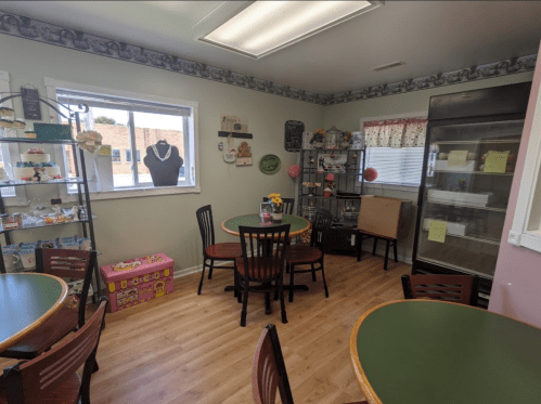 Cozy café interior with green tables, black chairs, a display shelf, and a cooler in a well-lit, inviting space.