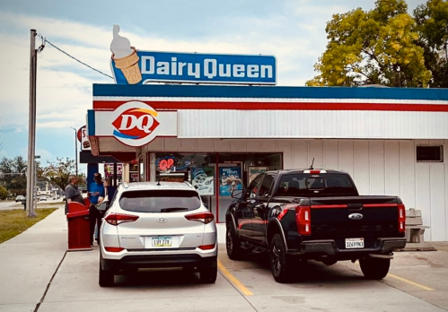 A Dairy Queen restaurant with two parked cars outside, featuring a sign with an ice cream cone.