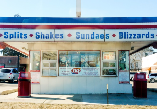 A Dairy Queen drive-thru with a menu featuring splits, shakes, sundaes, and Blizzards. Red trash bins in front.