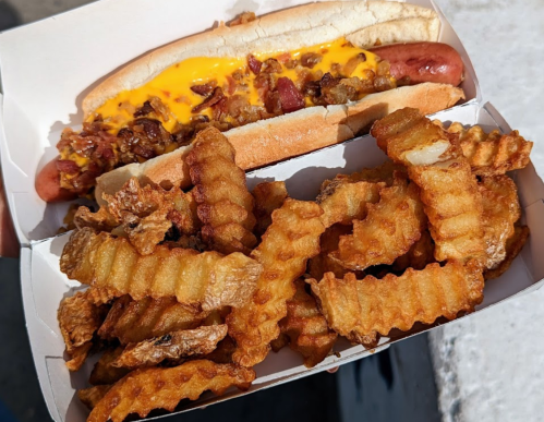 A hot dog topped with cheese and bacon, served with a side of crispy crinkle-cut fries in a takeout box.