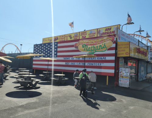 A colorful Nathan's restaurant with a large American flag mural, outdoor seating, and a carnival atmosphere.